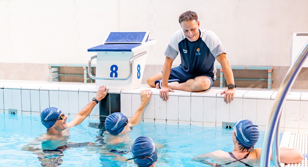 Trainer zit aan badrand en geeft vier zwemmers instructie